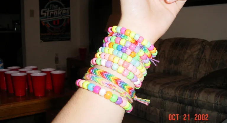 A lo-fi digicam photo from a house party in the early 2000s: a close-up of someone's wrist wearing a collection of those…