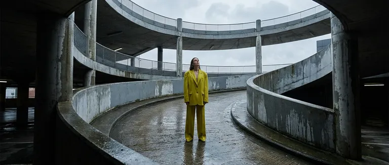 A wide editorial of a woman in a chartreuse yellow oversized blazer and coordinated trousers standing in a brutalist car…