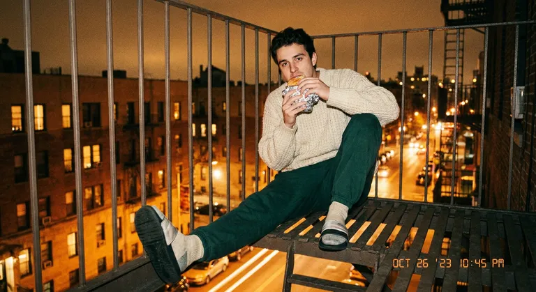 A lo-fi digicam shot of a guy sitting on a fire escape in New York at night, one leg dangling. He wears a heavy knit fis…