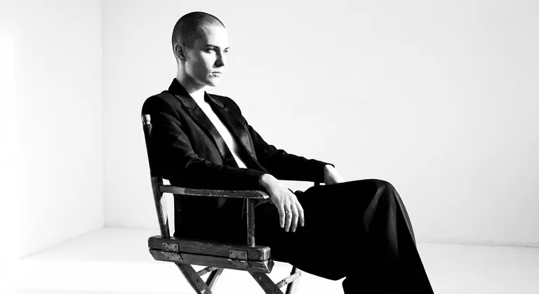 A high-contrast black and white editorial portrait of a non-binary model with a shaved head, sitting side-on in a wooden director's chair in an empty white studio. They wear a sharply tailored tuxedo jacket with nothing underneath, and wide-leg trousers. One hand rests on a knee. Strong single-source side lighting creates a dramatic split — half the face in bright highlight, half in deep shadow. Shot on a 85mm with film grain added in post. Mood is austere and commanding.
