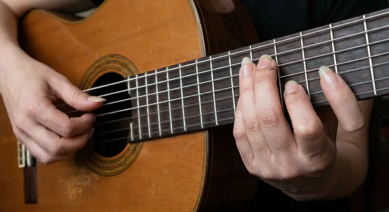 A close-up of a woman's fingers on the strings of a classical guitar — mid-chord, fingers pressed. The nails on the righ…