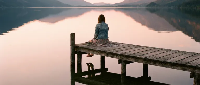 A wide shot at magic hour of a woman sitting alone on a wooden pier extending into a still lake, feet dangling over the edge, shoes beside her. She faces away from the camera. She wears a worn denim jacket over a floral dress, the hem trailing slightly. The lake is glassy and reflects the pink-gold sky exactly. The pier planks are old and weathered. The image is entirely about solitude, reflection, and that specific quality of late afternoon quiet.