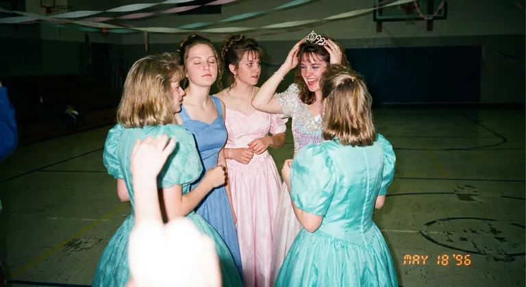A lo-fi film snapshot of a group of girls at prom: the kind of picture where someone's eyes are closed, someone else is looking away, the lighting from the camera flash makes everyone slightly alien. They wear a range of gowns — a few matching accidents. One girl is tilting her tiara back onto her head mid-photo. The linoleum gym floor is visible below. The photo is imperfect in every way and perfect because of it.
