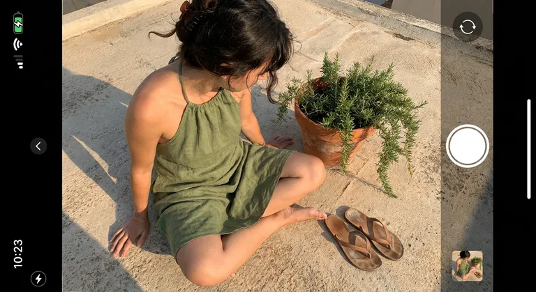 A candid overhead iPhone shot of a woman sitting cross-legged on a sun-bleached concrete rooftop, a terracotta pot of trailing rosemary beside her. She wears a moss-green linen halter dress, sand-dusted flip-flops discarded nearby. Her hair is pinned loosely, a few dark strands escaping over her shoulder. Late afternoon light rakes across the concrete, casting long golden shadows. The color palette is warm ochre and sage. Texture-forward: rough concrete grain, wrinkled linen, dried herb needles in sharp focus.