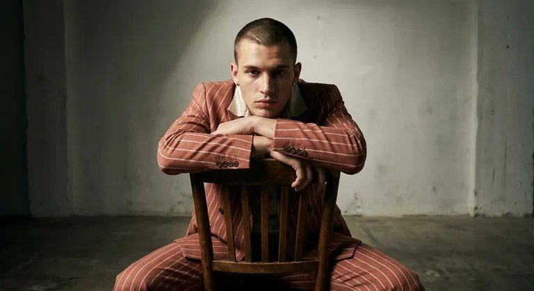 A fashion editorial shot of a male model, early 20s, sitting backwards on a chair in a sparse studio, arms folded across…